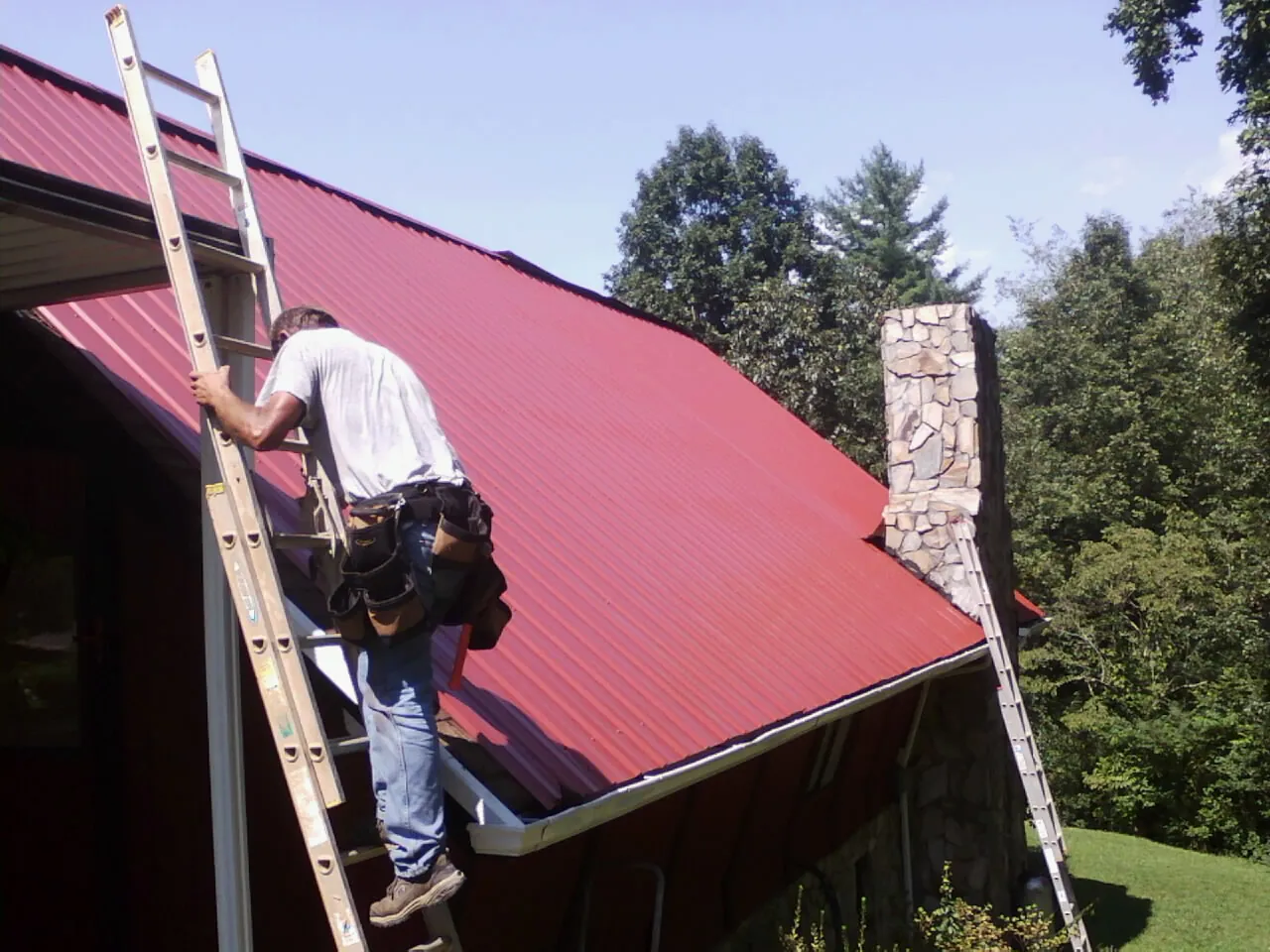 Roofer on a ladder beside a red metal roof and stone chimney — Soffit & Fascia Repair by Roofing Brothers in Ann Arbor